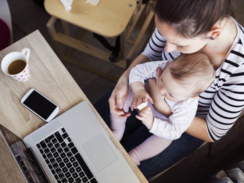 Bird's eye view of a working mother holding a baby and looking at a laptop screen